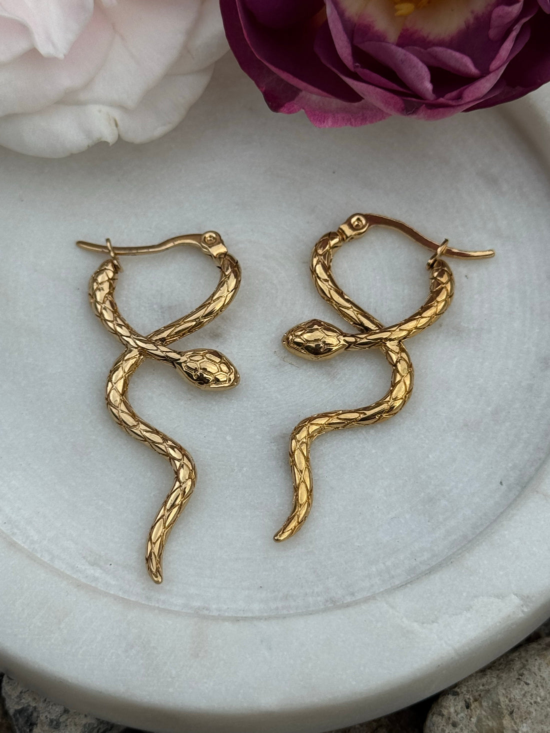 Gold snake-shaped hoop earrings with a detailed textured scale design, displayed on a round white stone surface with soft pink and purple roses in the background.