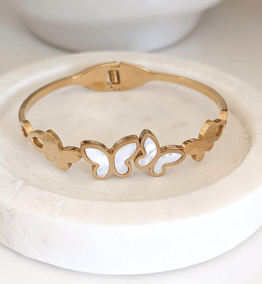 Gold butterfly cuff bracelet with mother-of-pearl inlay, displayed on a textured surface with floral décor. 18k gold plated, waterproof and tarnish-free jewellery.