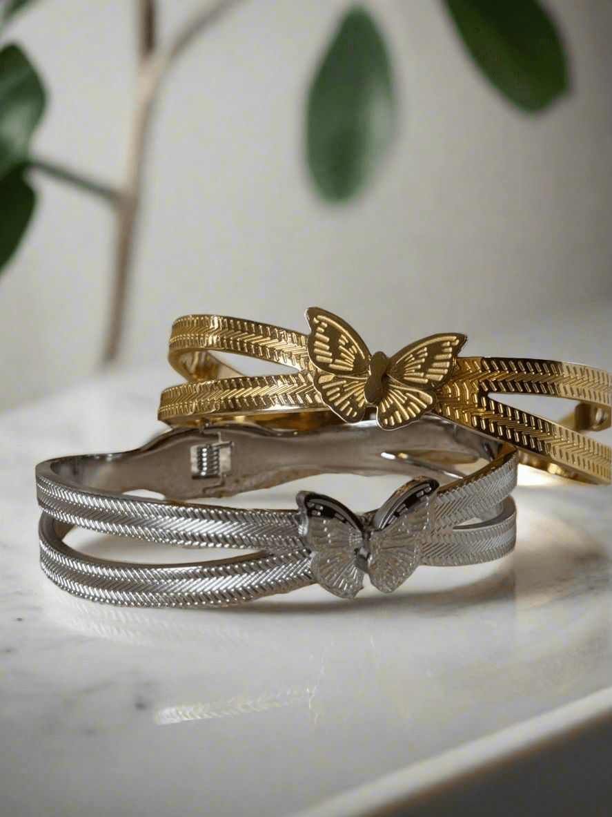 Two textured metal bracelets with butterfly clasps—one in silver and the other in gold—displayed on a cream-colored stone surface with a white ceramic lotus and an orange flower in the background.
