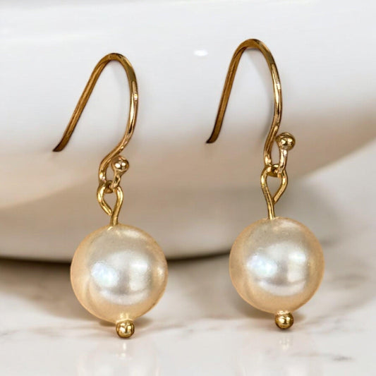 Elegant 14K gold-filled drop earrings featuring genuine Swarovski pearls, hanging on a gold jewelry stand with soft, natural lighting.