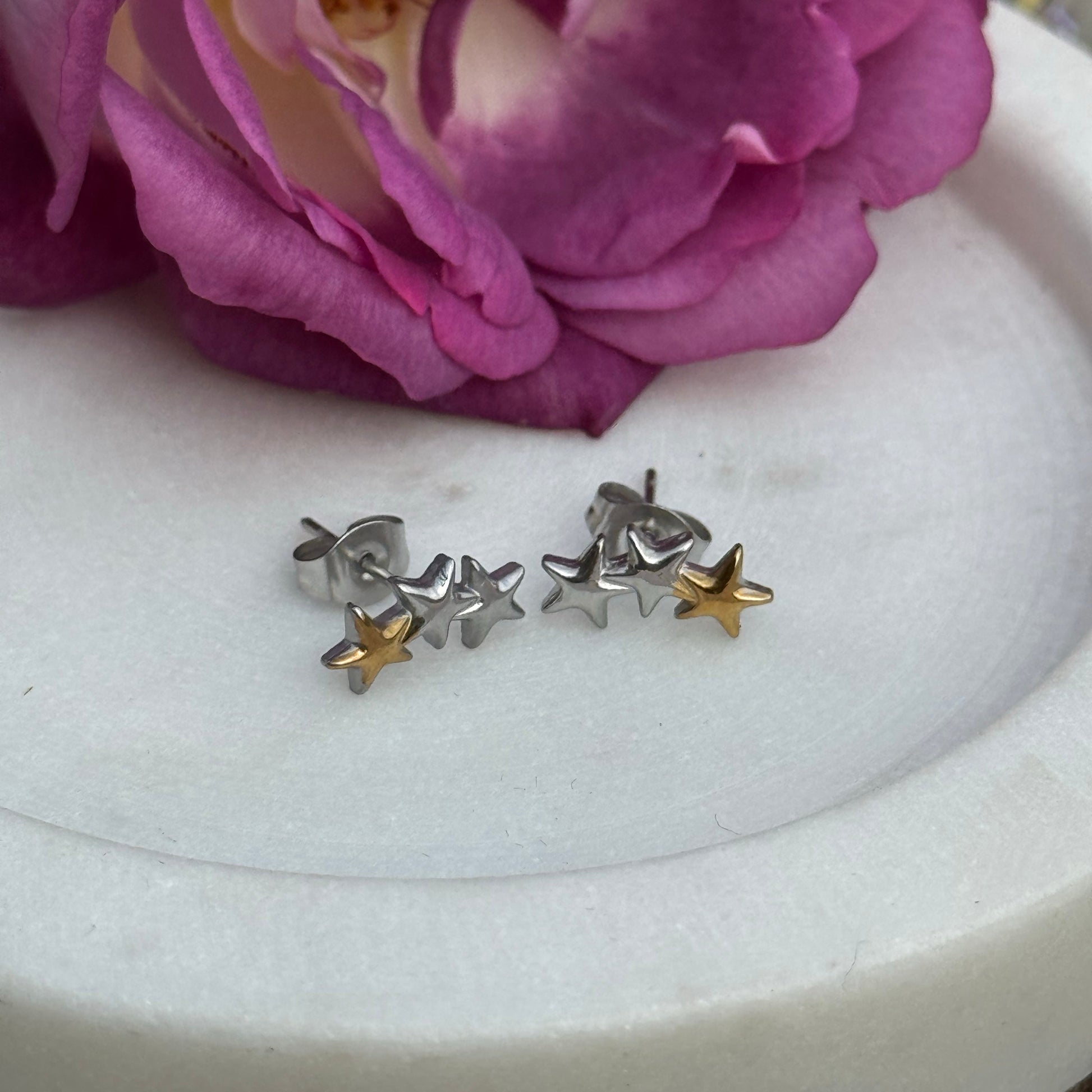 Two-tone stainless steel triple star stud earrings with silver and gold stars, displayed on a white marble tray with a purple rose
