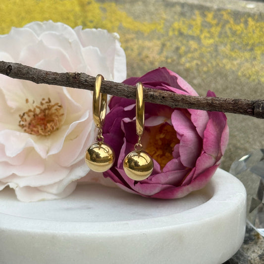 Gold ball drop hoop earrings made of stainless steel with 18K PVD gold plating, hanging from a small branch amongst a purple rose