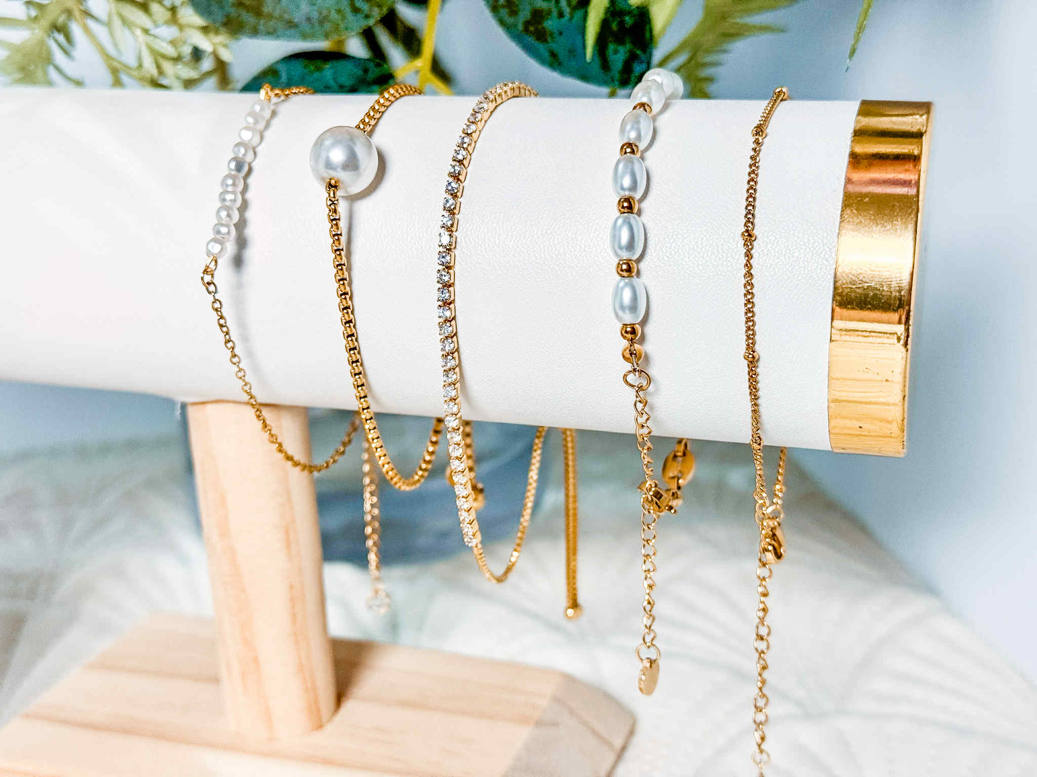 Set of gold and pearl bracelets on a white cylindrical stand with a blurred green plant background.