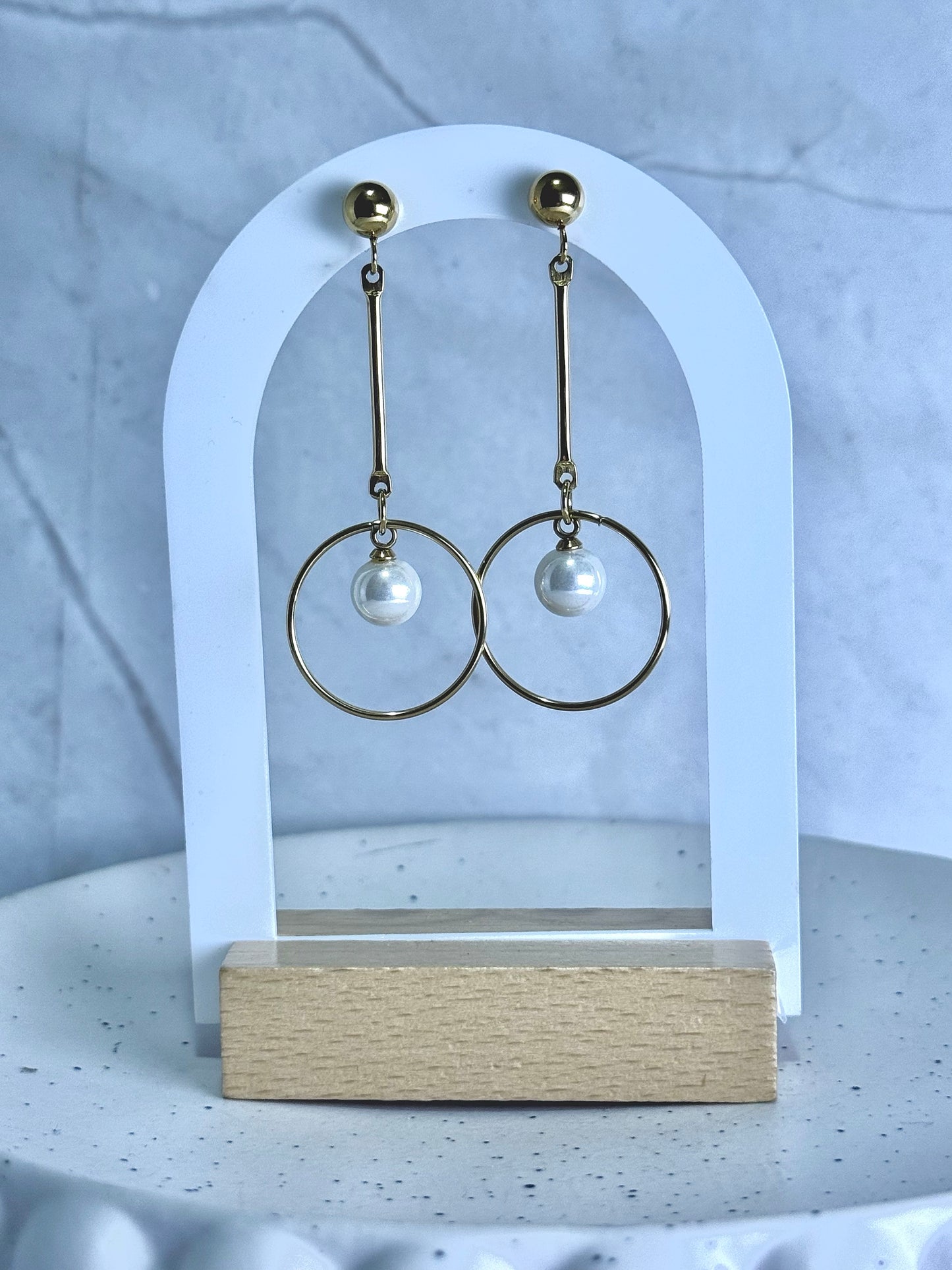 Gold hoop earrings with pearl drops on a white arch stand against a light gray background.