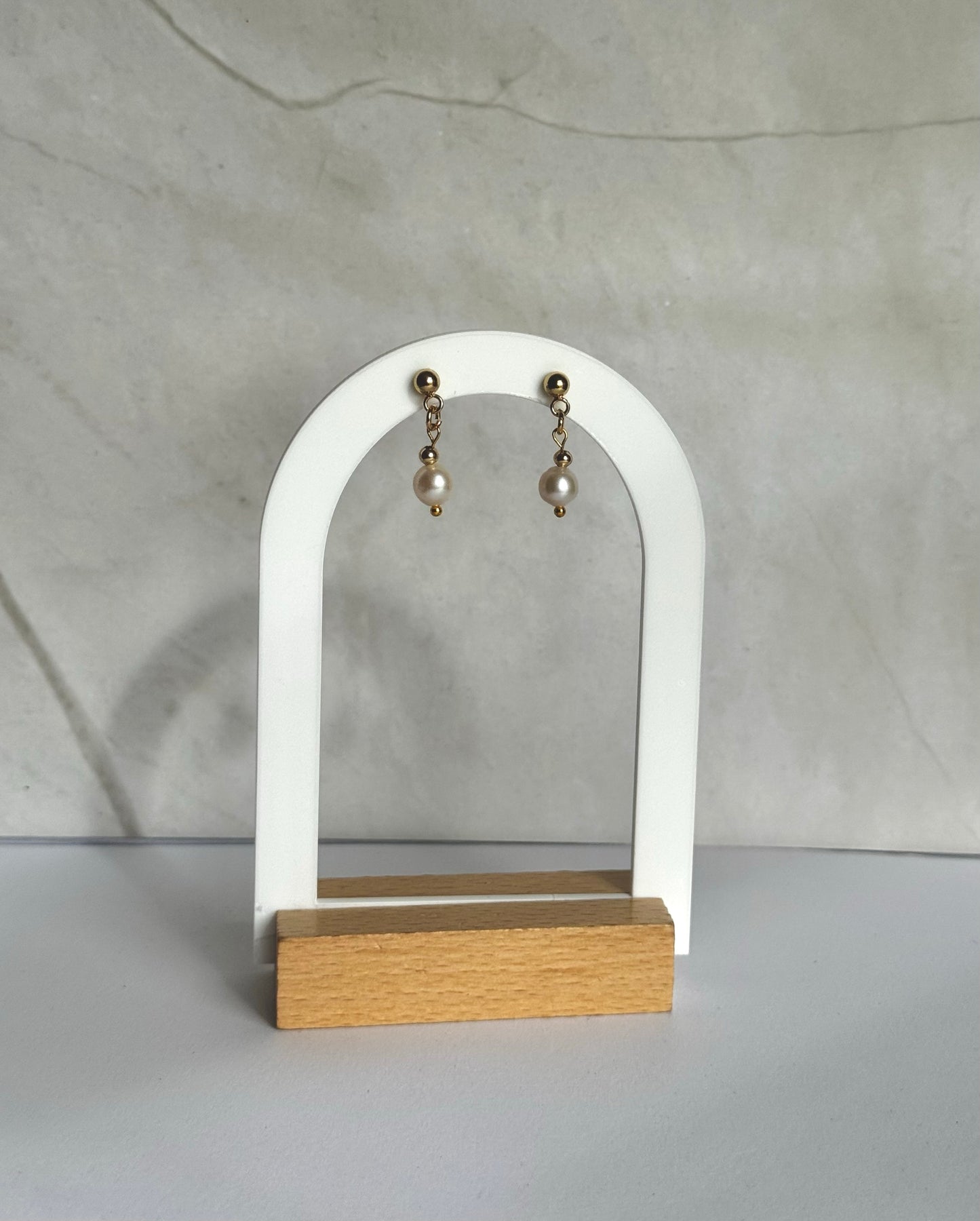 Pair of pearl earrings on a white arch-shaped stand with a wooden base against a marble background.