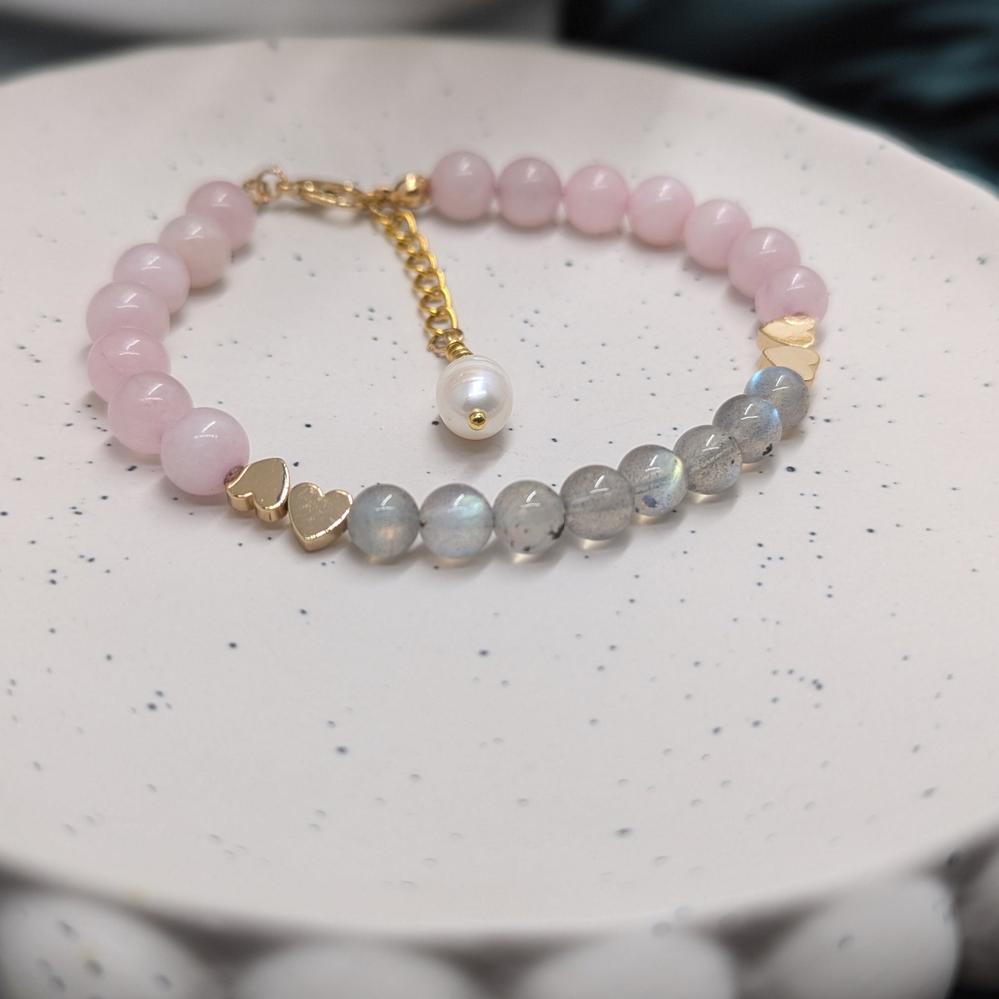 Handmade beaded bracelet featuring natural rose quartz and labradorite gemstones with a freshwater pearl charm, gold-plated heart spacers, and extender chain, displayed on a ceramic dish.