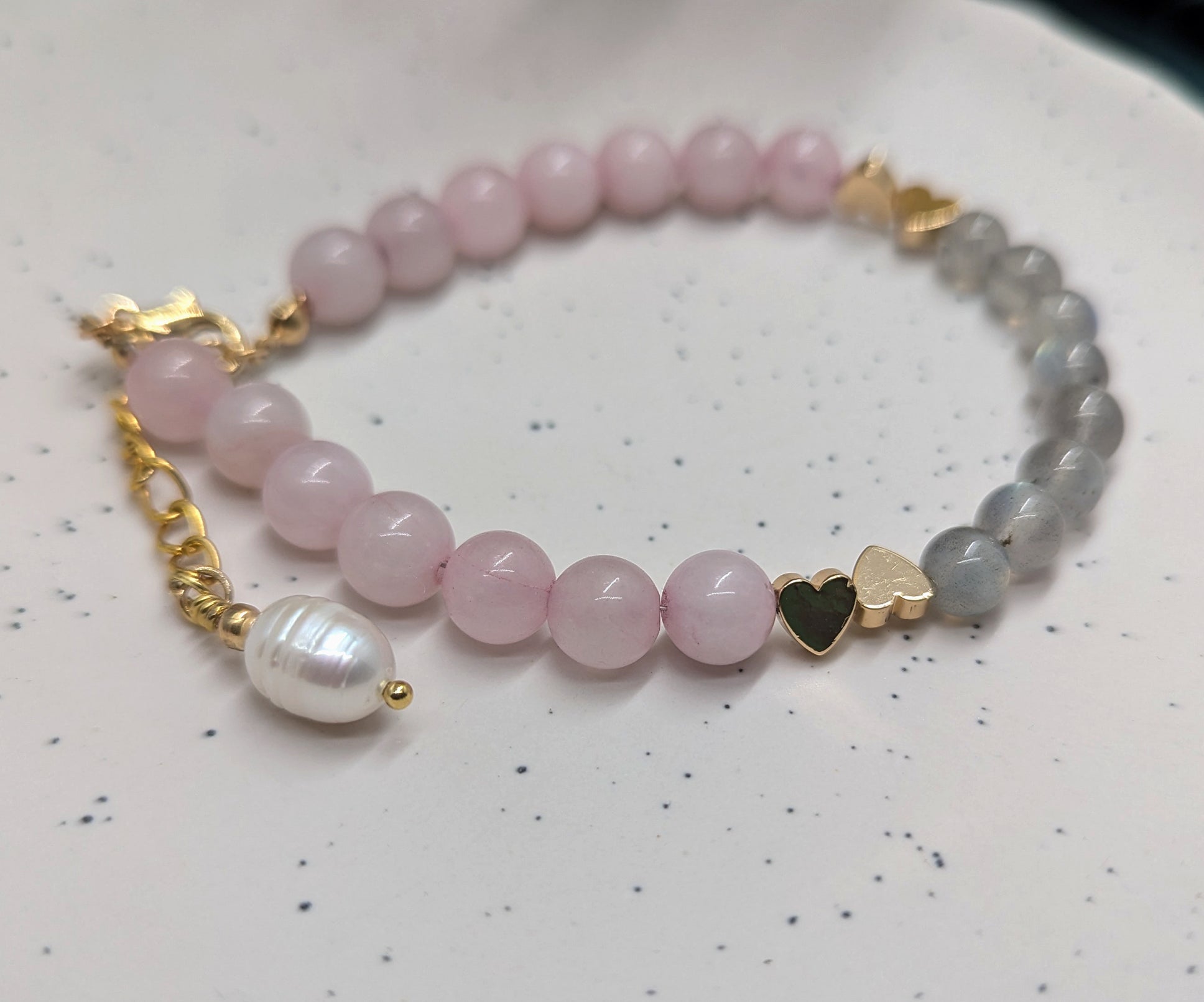 Close-up of a handmade bracelet featuring round rose quartz and labradorite beads, accented with gold heart spacers and a freshwater pearl charm on a gold extender chain,