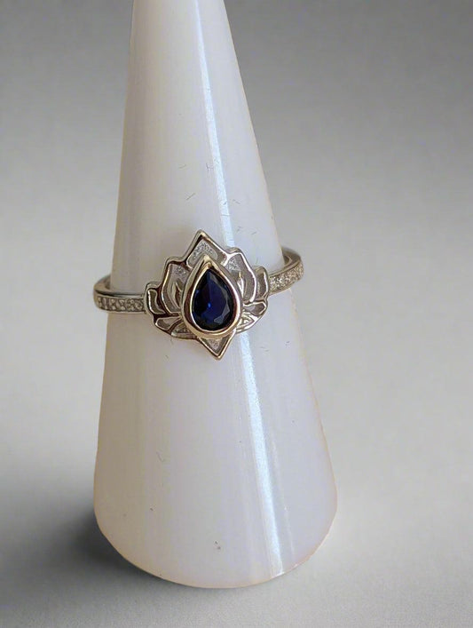 A sterling silver adjustable ring featuring a teardrop-shaped deep blue sapphire set in a delicate lotus-inspired design. The band is adorned with pave-set cubic zirconia accents, adding a touch of sparkle. Perfect for everyday wear or special occasions.