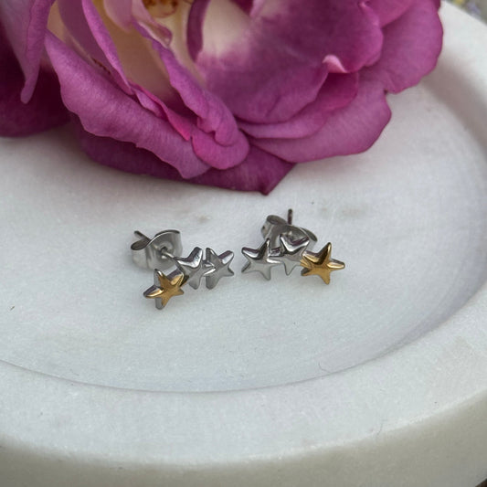 Two-tone stainless steel triple star stud earrings with silver and gold stars, displayed on a white marble tray with a purple rose