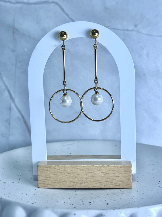 Gold hoop earrings with pearl drops on a white arch stand against a light gray background.