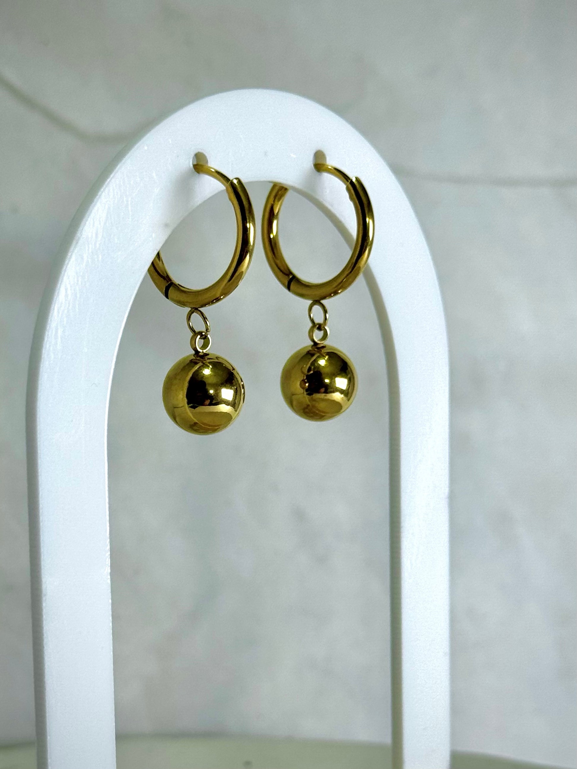 Gold hoop earrings with spherical charms on a white stand against a white background