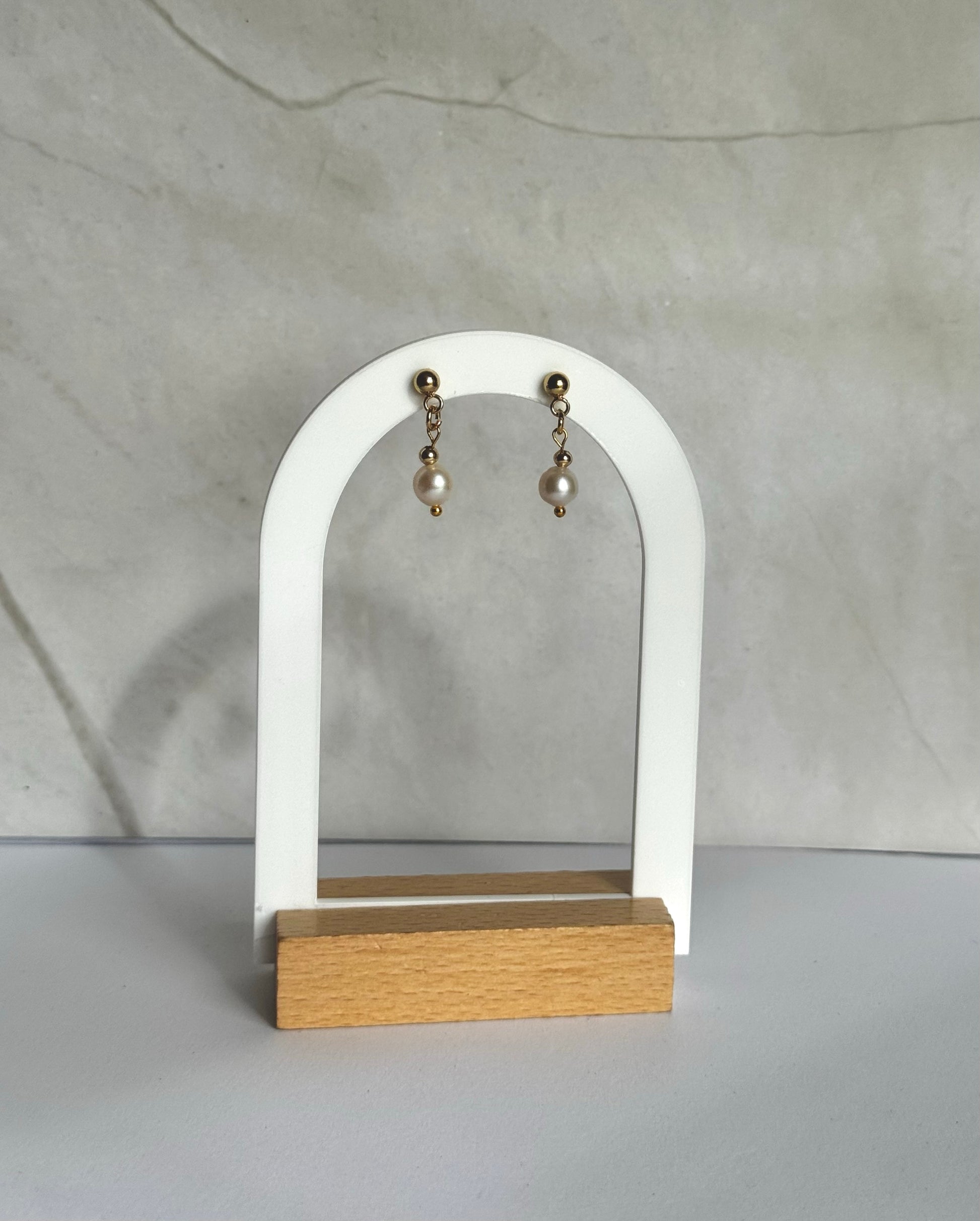 Pair of pearl earrings on a white arch-shaped stand with a wooden base against a marble background.